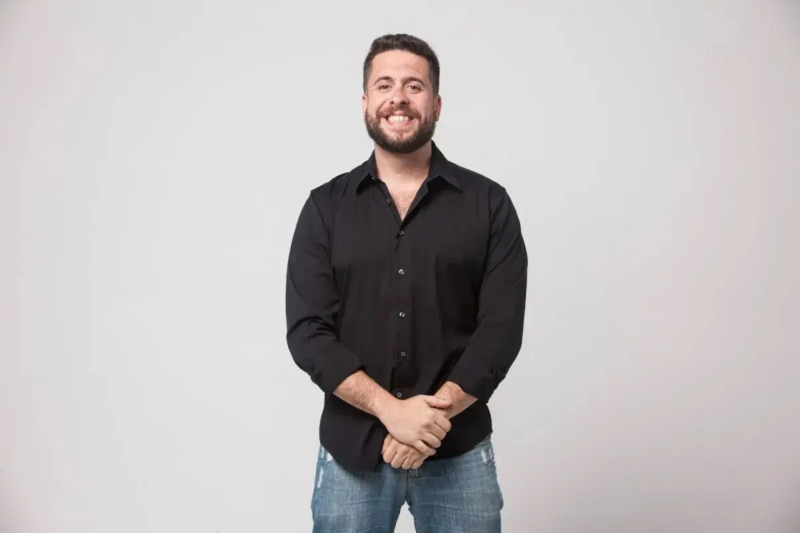 Humorista posa sorrindo, usando camisa preta e calça jeans, diante de um fundo claro