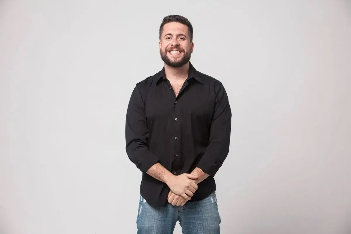 Humorista posa sorrindo, usando camisa preta e calça jeans, diante de um fundo claro