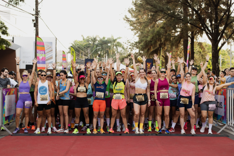 Grupo de mulheres com roupas esportivas e números de peito levanta os braços antes da largada de uma corrida de rua.