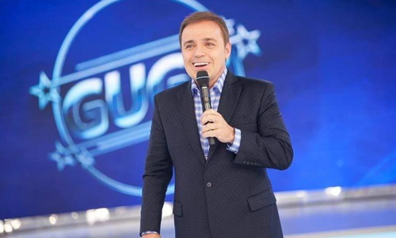 Apresentador de televisão sorrindo e segurando um microfone em um palco com cenário azul ao fundo.