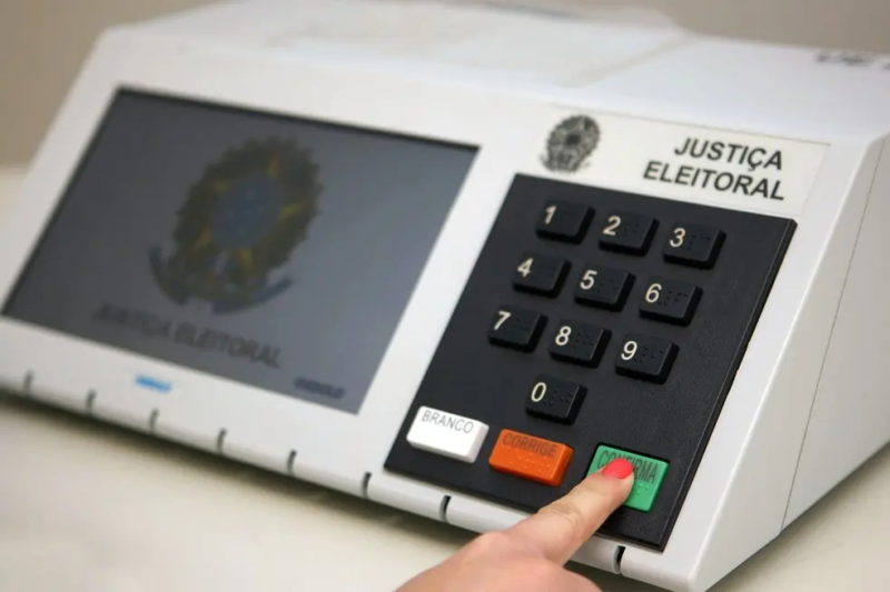 Close de uma urna eletrônica brasileira enquanto uma pessoa pressiona a tecla verde “Confirma”.