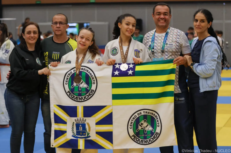 Duas judocas de Anápolis exibem medalhas e seguram bandeiras ao lado de integrantes da equipe do Colégio Pequenuchos Teens nos Jogos Escolares Brasileiros.