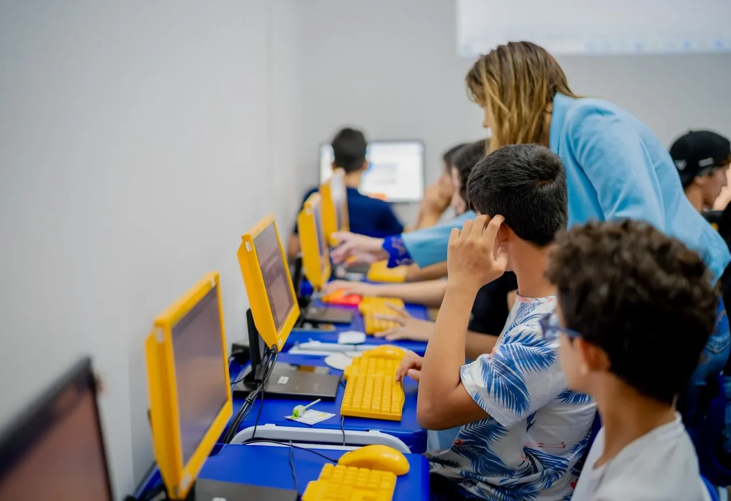 Alunos utilizam computadores com monitores e teclados amarelos enquanto uma mulher orienta uma atividade em sala de informática.