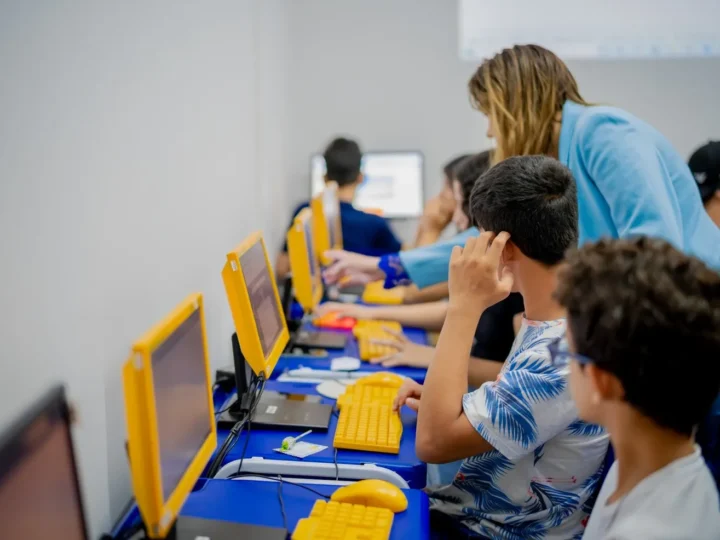 Alunos utilizam computadores com monitores e teclados amarelos enquanto uma mulher orienta uma atividade em sala de informática.
