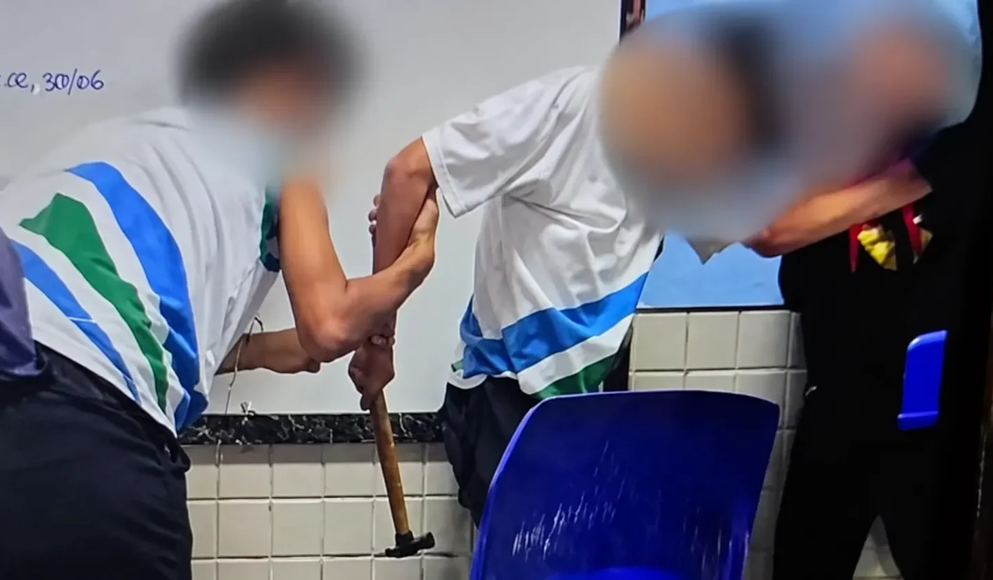 Pessoas com rostos desfocados em uma sala de aula, com um martelo visível próximo a uma cadeira azul.