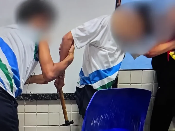 Pessoas com rostos desfocados em uma sala de aula, com um martelo visível próximo a uma cadeira azul.