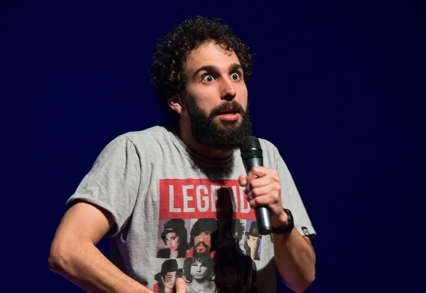 Murilo Couto segura um microfone durante apresentação, usando camiseta cinza diante de um fundo azul.