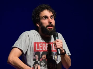 Murilo Couto segura um microfone durante apresentação, usando camiseta cinza diante de um fundo azul.