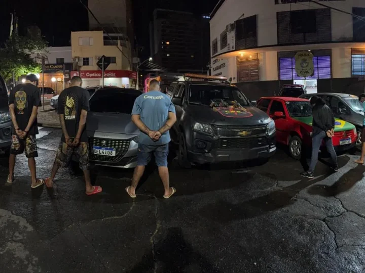 Cinco homens de costas diante de carros e viaturas policiais, à noite, em frente a uma unidade da Polícia Civil.