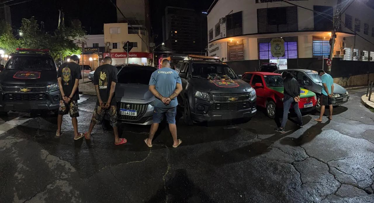Cinco homens de costas diante de carros e viaturas policiais, à noite, em frente a uma unidade da Polícia Civil.