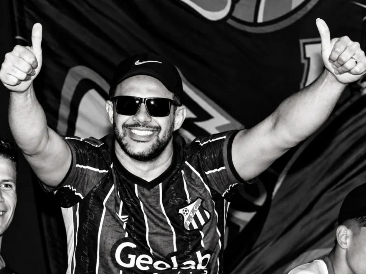 Rodrigo Costa Teixeira sorri com os braços levantados e os polegares para cima, usando boné, óculos escuros e camisa do Anápolis Futebol Clube.