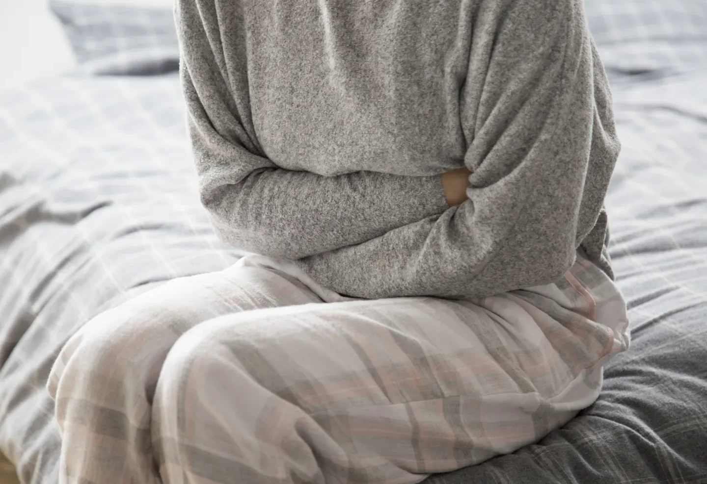 Pessoa sentada na cama, com blusa cinza e calça de pijama, pressionando os braços contra o abdômen.