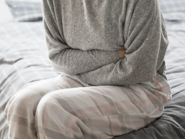 Pessoa sentada na cama, com blusa cinza e calça de pijama, pressionando os braços contra o abdômen.