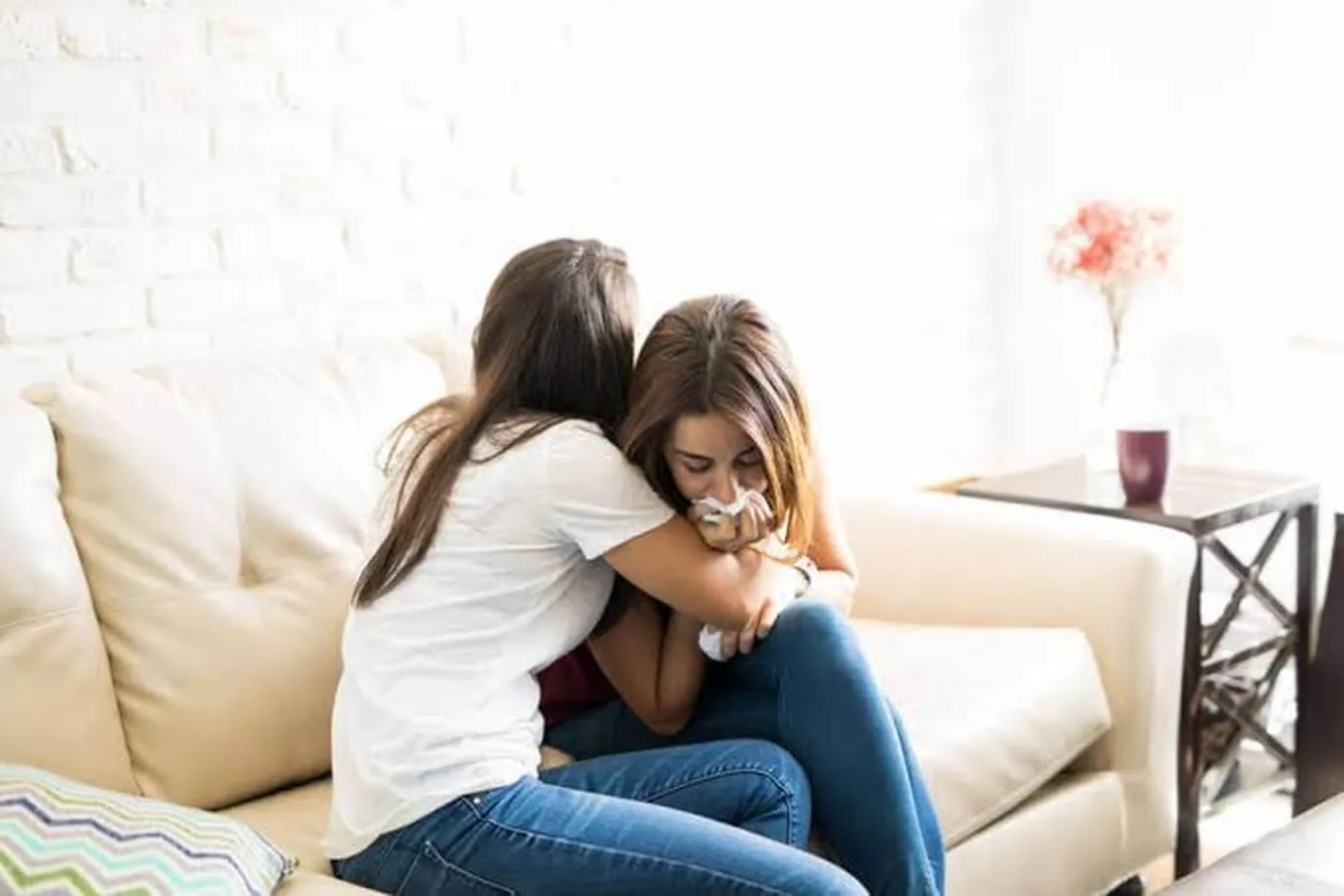Duas mulheres sentadas em um sofá; uma abraça a outra, que mantém a cabeça baixa e segura um lenço próximo ao rosto