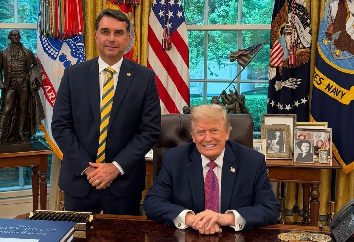 Flávio Bolsonaro em pé ao lado de Donald Trump, sentado atrás de uma mesa, com bandeiras dos Estados Unidos ao fundo.