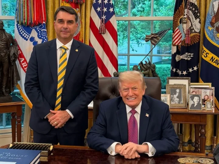 Flávio Bolsonaro em pé ao lado de Donald Trump, sentado atrás de uma mesa, com bandeiras dos Estados Unidos ao fundo.