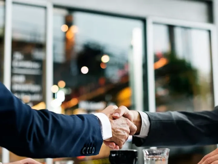 Dois homens de terno apertam as mãos sobre uma mesa, com copos e uma xícara.