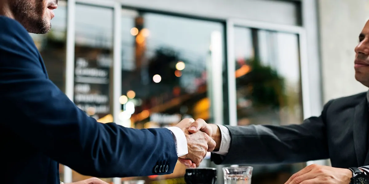 Dois homens de terno apertam as mãos sobre uma mesa, com copos e uma xícara.