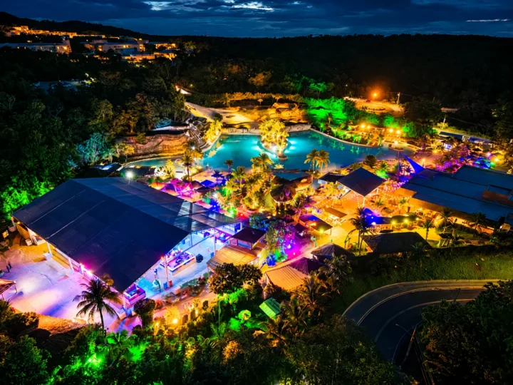 Vista aérea noturna do Hot Park, com piscinas, tendas e áreas de eventos iluminadas por luzes coloridas.