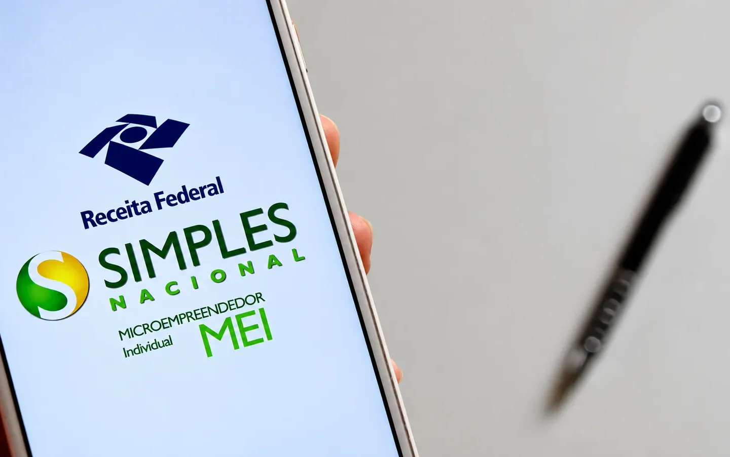 Celular exibe os logotipos da Receita Federal, do Simples Nacional e do MEI, com uma caneta desfocada ao fundo.