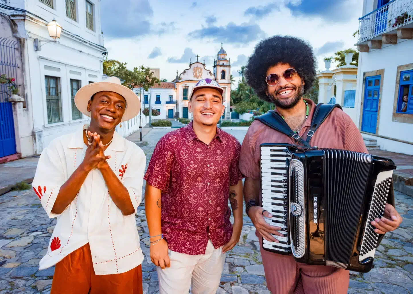 Jota.pê, João Gomes e Mestrinho posam sorrindo em uma rua de pedras, com Mestrinho segurando uma sanfona.