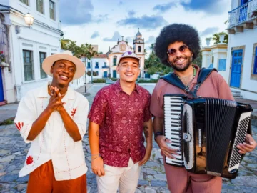 Jota.pê, João Gomes e Mestrinho posam sorrindo em uma rua de pedras, com Mestrinho segurando uma sanfona.