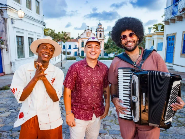 Jota.pê, João Gomes e Mestrinho posam sorrindo em uma rua de pedras, com Mestrinho segurando uma sanfona.