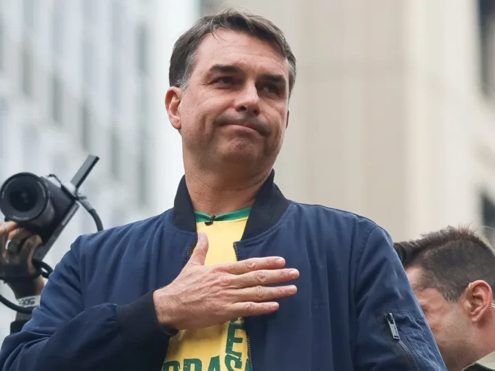 Flávio Bolsonaro com a mão sobre o peito, usando jaqueta azul e camiseta amarela, em ambiente externo.