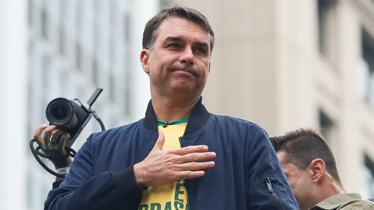 Flávio Bolsonaro com a mão sobre o peito, usando jaqueta azul e camiseta amarela, em ambiente externo.