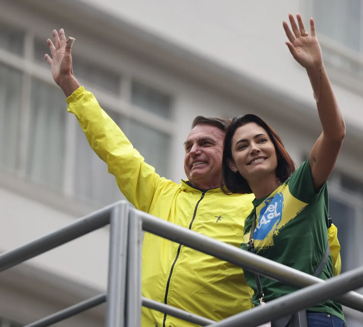 Jair e Michelle Bolsonaro sorriem e acenam em uma plataforma, usando roupas nas cores amarela e verde.