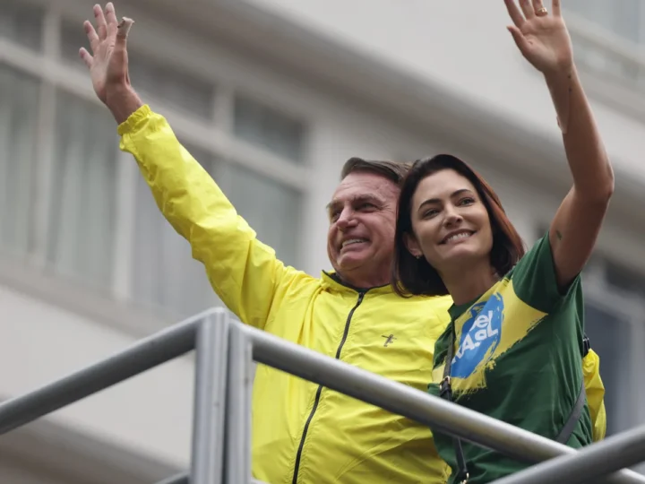 Jair e Michelle Bolsonaro sorriem e acenam em uma plataforma, usando roupas nas cores amarela e verde.