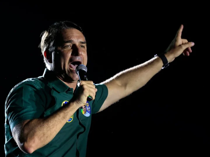 Flávio Bolsonaro com camisa verde, segurando um microfone e apontando para cima com o braço erguido.