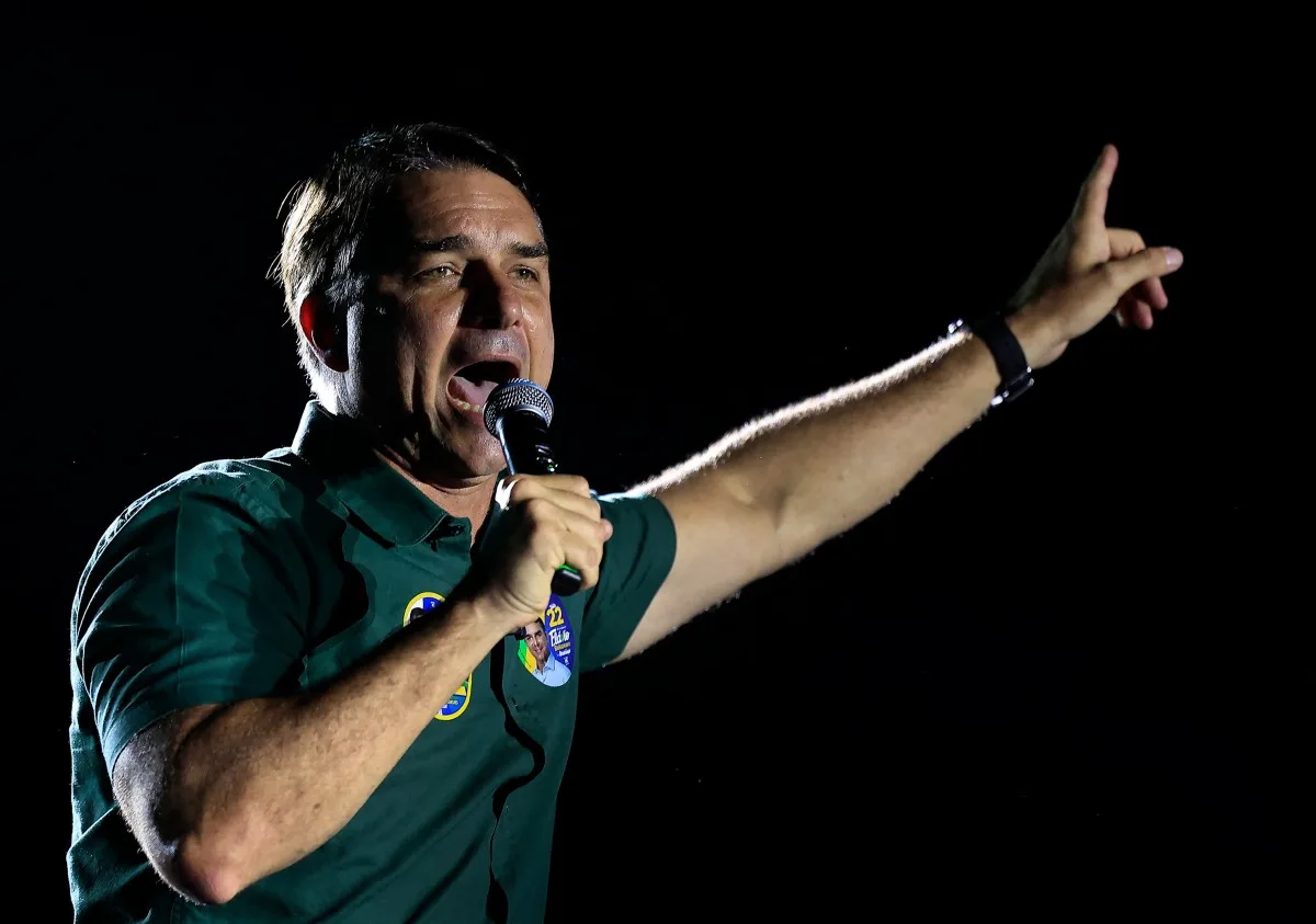Flávio Bolsonaro com camisa verde, segurando um microfone e apontando para cima com o braço erguido.
