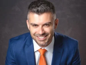 Homem sorrindo, com terno azul, camisa branca e gravata laranja, diante de fundo cinza.