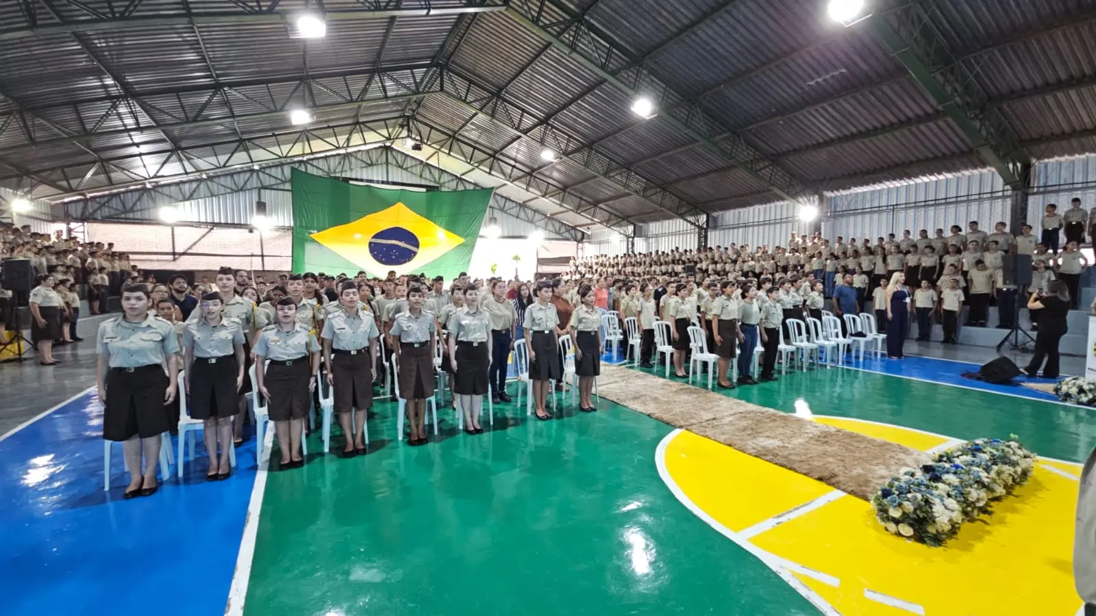 Estudantes uniformizados reunidos em uma quadra coberta, com uma grande bandeira do Brasil ao fundo.
