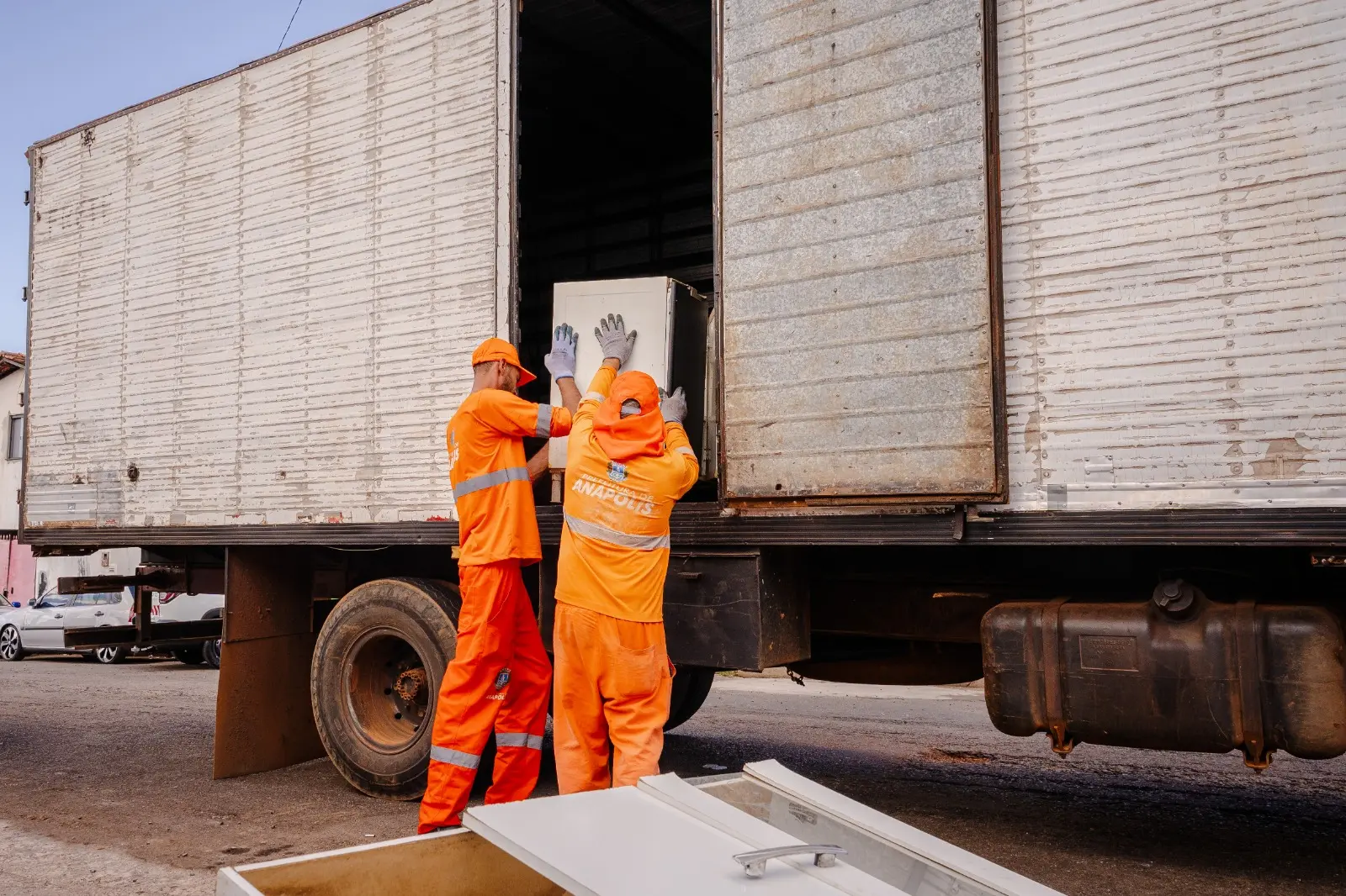 Dois trabalhadores com uniformes laranja carregam um móvel para dentro de um caminhão.